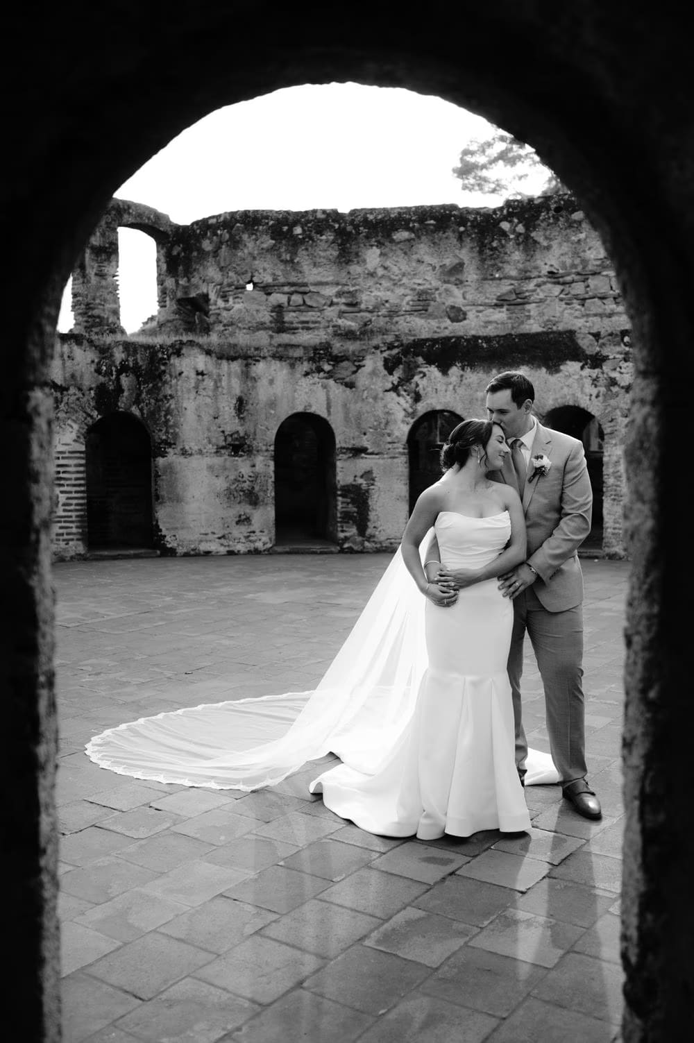 Black-and-white portrait of bride and groom embracing inside a stone archway with the ruined courtyard glowing behind them.