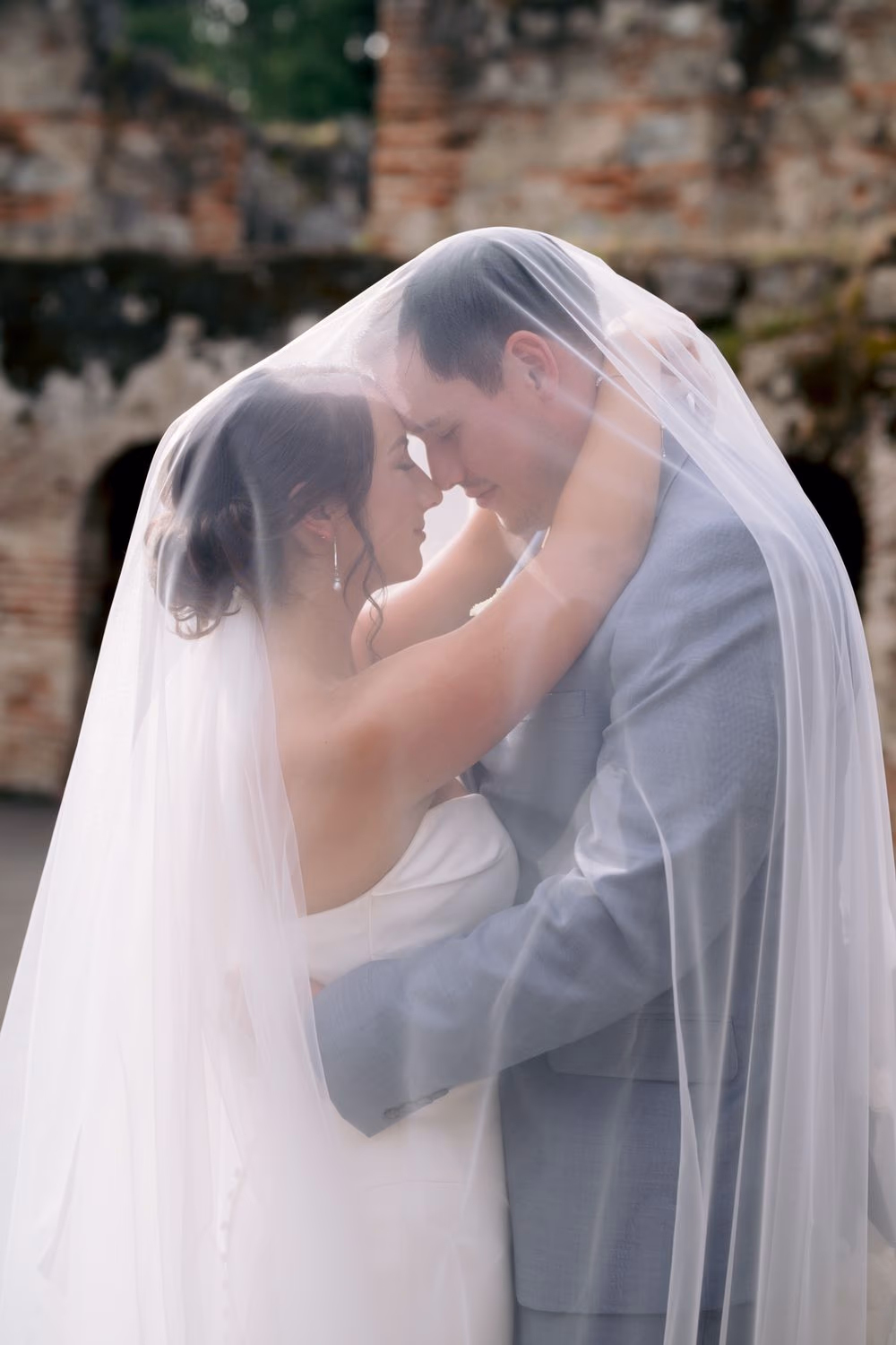 Bride and groom press foreheads together beneath her cathedral veil, brick ruins blurred behind them.