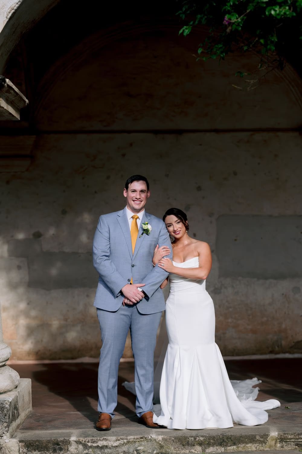 Bride and groom pose under a colonial arch in soft afternoon light, the bride leaning into him.