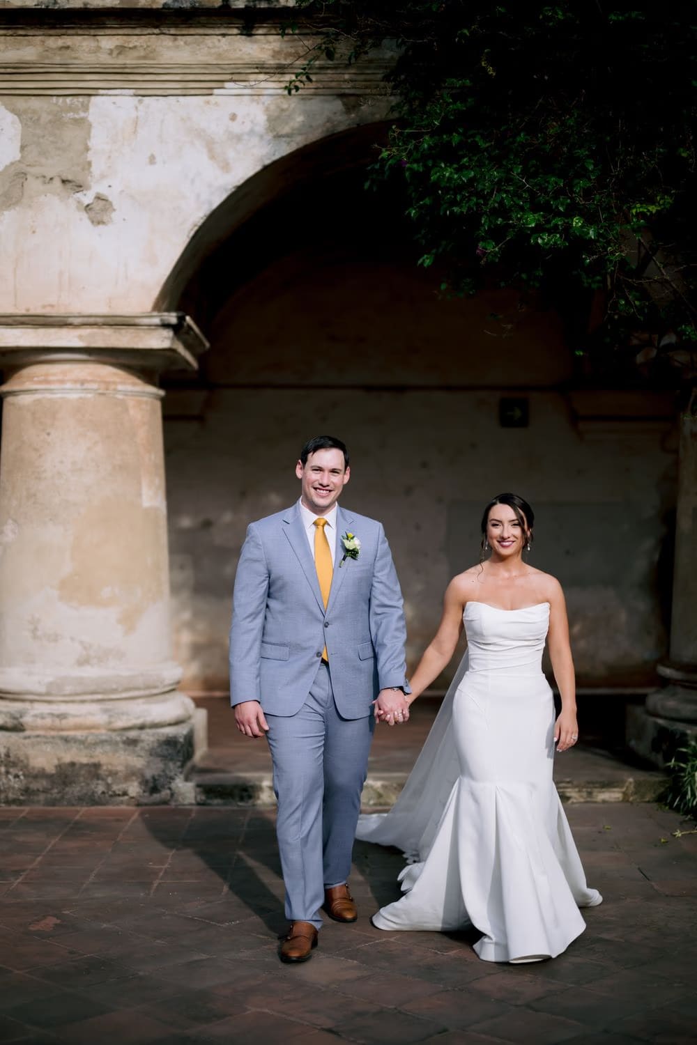 Newlyweds walk hand in hand beneath a tall colonial archway draped with bougainvillea, both smiling at the camera.