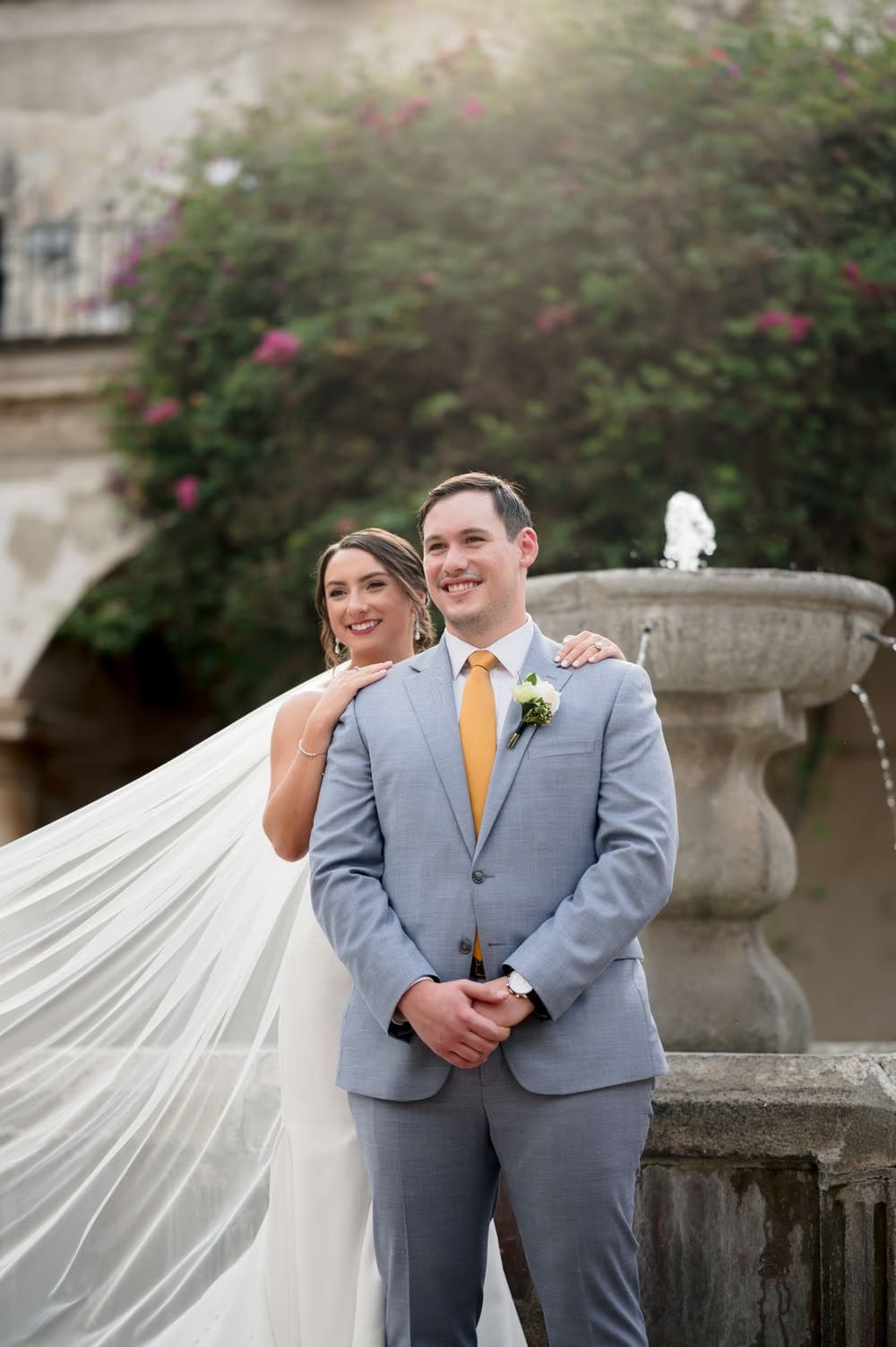 Bride's veil flies sideways as the couple poses by a colonial fountain framed by bougainvillea in golden afternoon light.