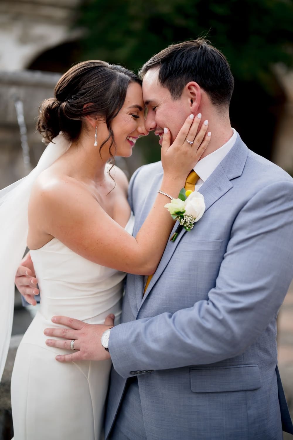 Bride cradles the groom's face in her hand, both laughing forehead to forehead as the fountain mists behind them.