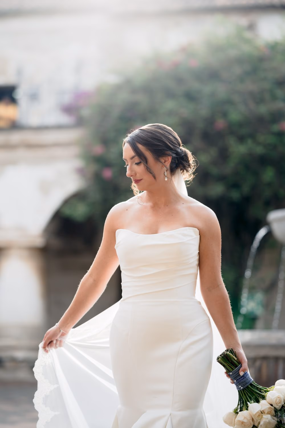 Solo bridal portrait — Alexis sweeps the train of her gown beside a bougainvillea-covered fountain in soft late light.