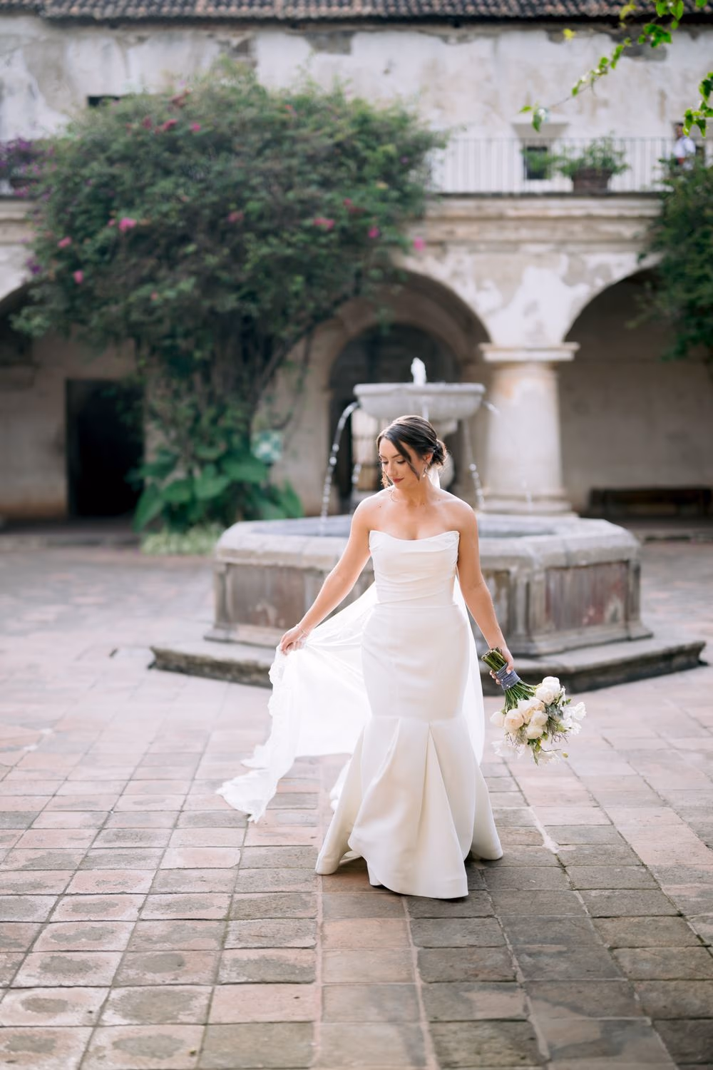 Bride walks across the cloister tiles holding her bouquet, a stone fountain glowing softly behind her.
