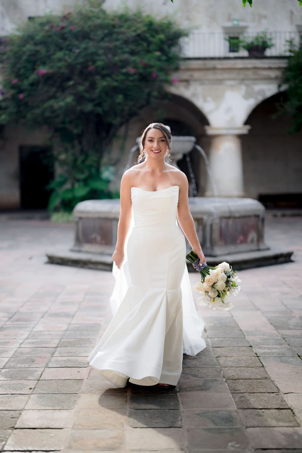 Bride smiles walking through the cloister courtyard, fountain and stone arches softly out of focus behind her.