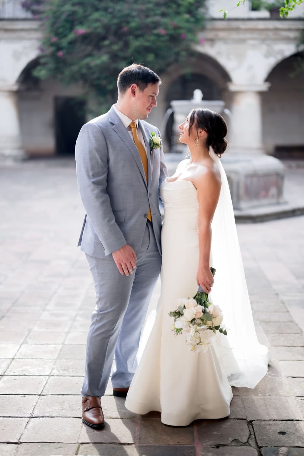 Bride looks up at her groom in the cloister courtyard, her veil draping behind her and his boutonniere catching the warm light.