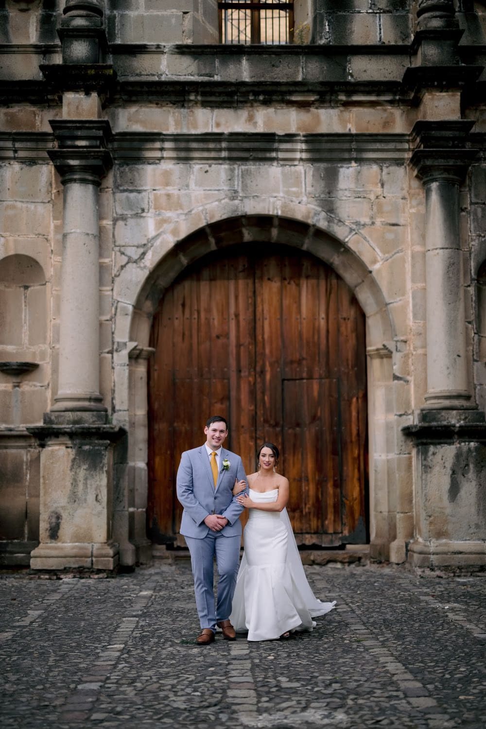 Alexis and Victor stand together in front of a massive arched wooden door at the convent entrance — formal couple portrait.