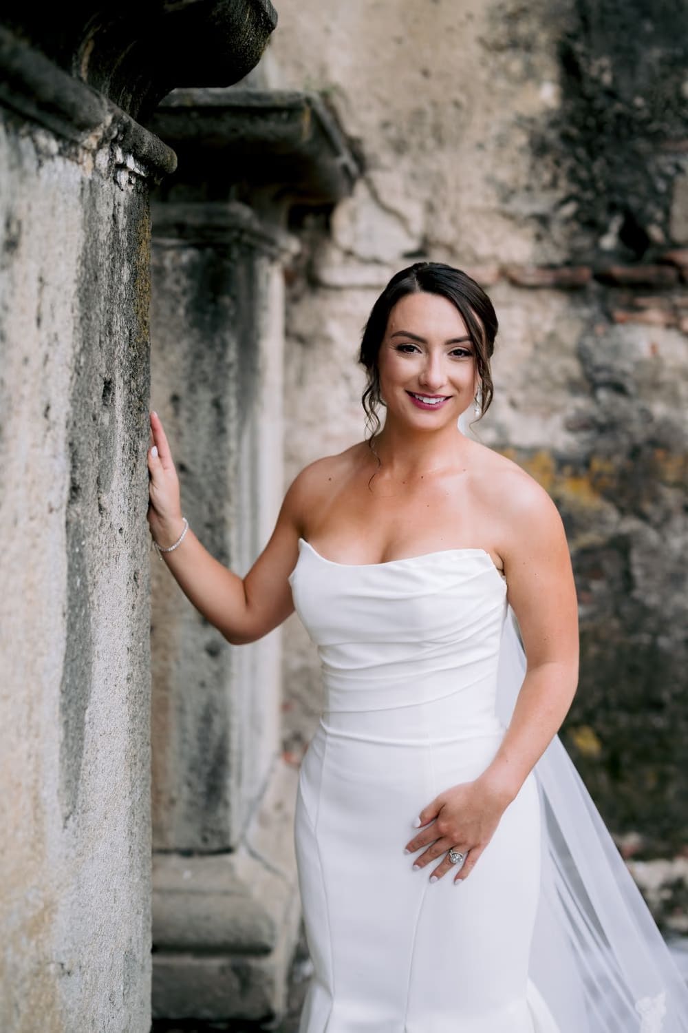 Bride smiles brightly beside a moss-touched stone column, late-afternoon glow lighting her face and engagement ring.