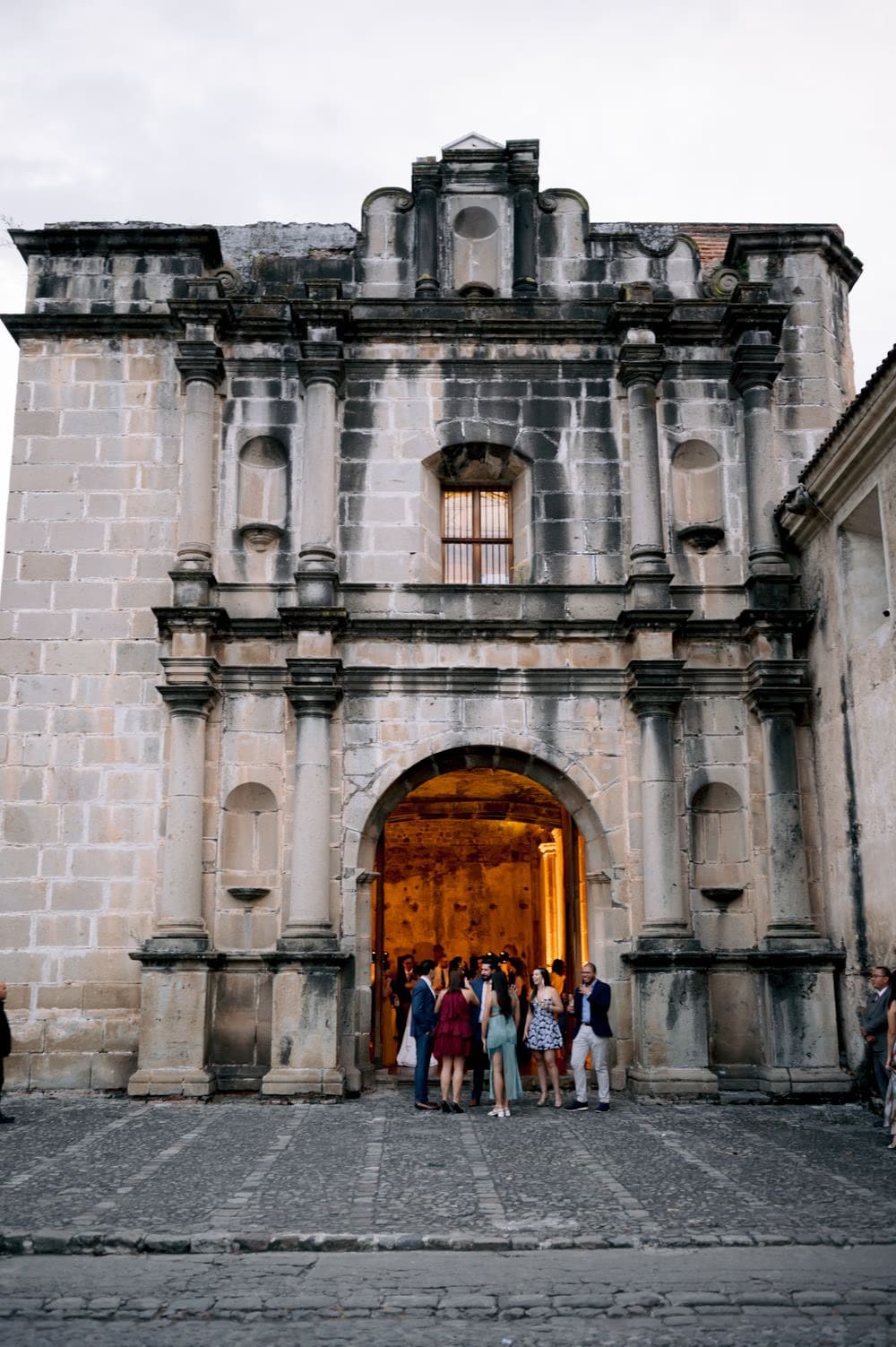 Guests gather inside the warmly lit arched entrance of the convent facade as twilight falls over the cobblestones outside.