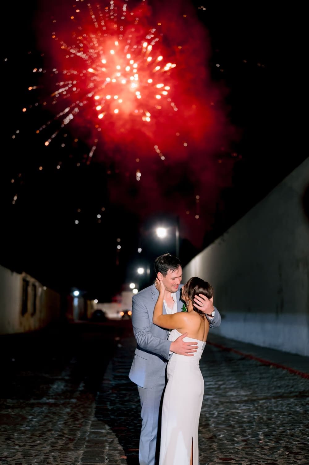 Bride and groom embrace on a cobblestone street as red fireworks burst above the rooftops at night in Antigua Guatemala.
