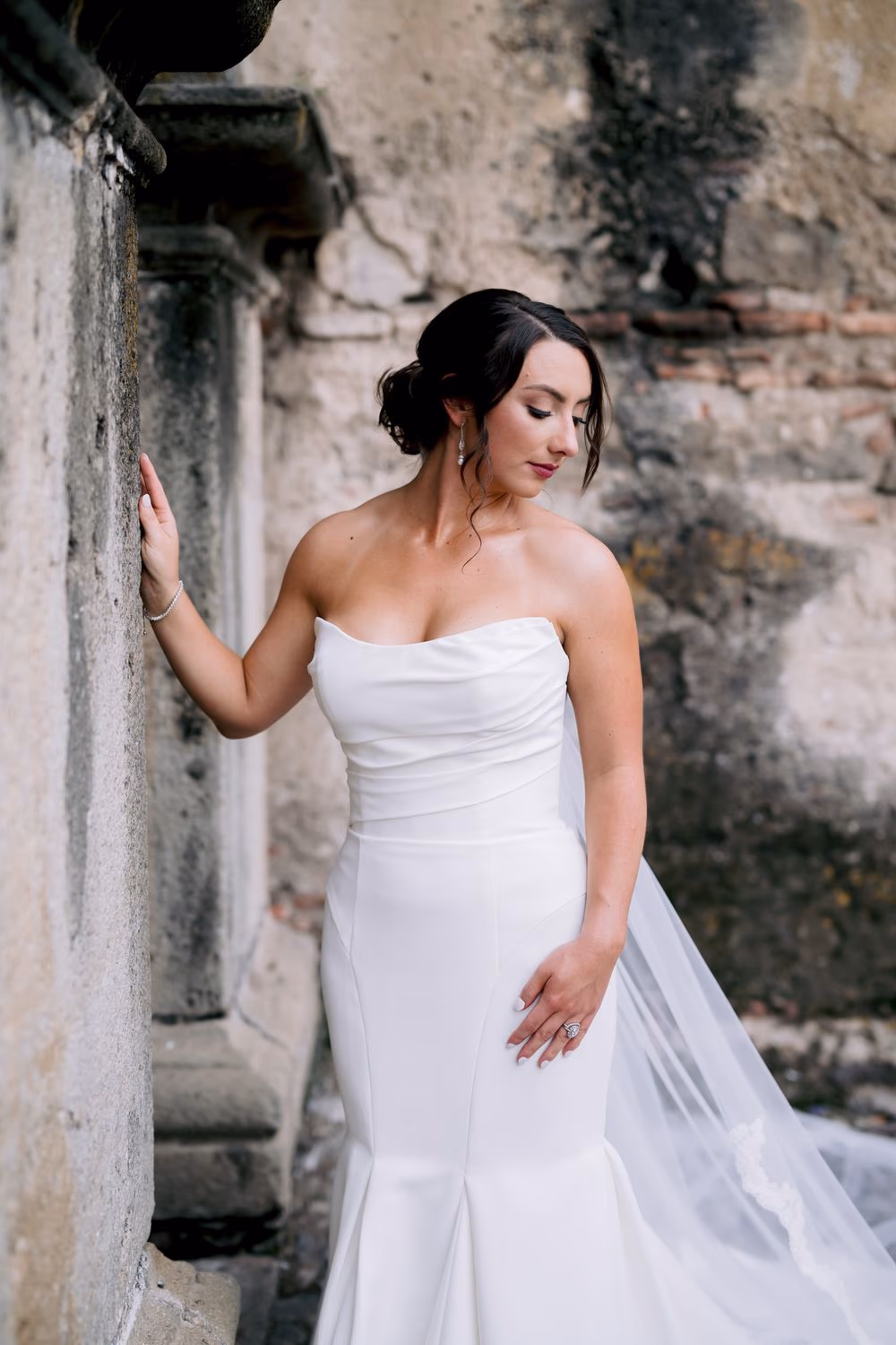 Bride Alexis leans against a weathered stone column in a strapless mermaid gown, golden afternoon light hitting the lime-mottled walls of Capuchinas Ruins, Antigua Guatemala — editorial bridal portrait.