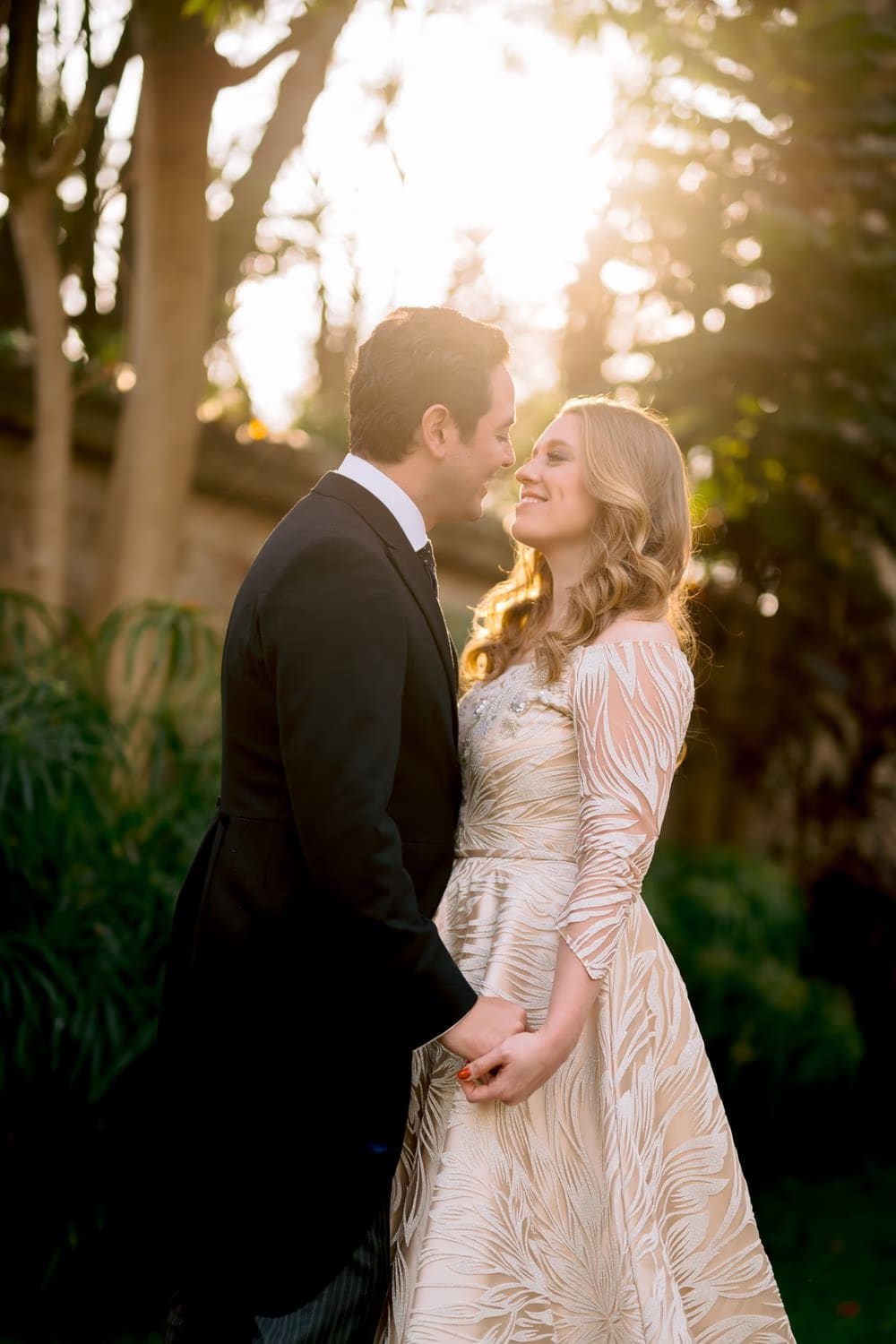 Bride and groom hold hands and lean in to share a smile, backlit by warm sunset light through the garden trees at their colonial Antigua wedding.