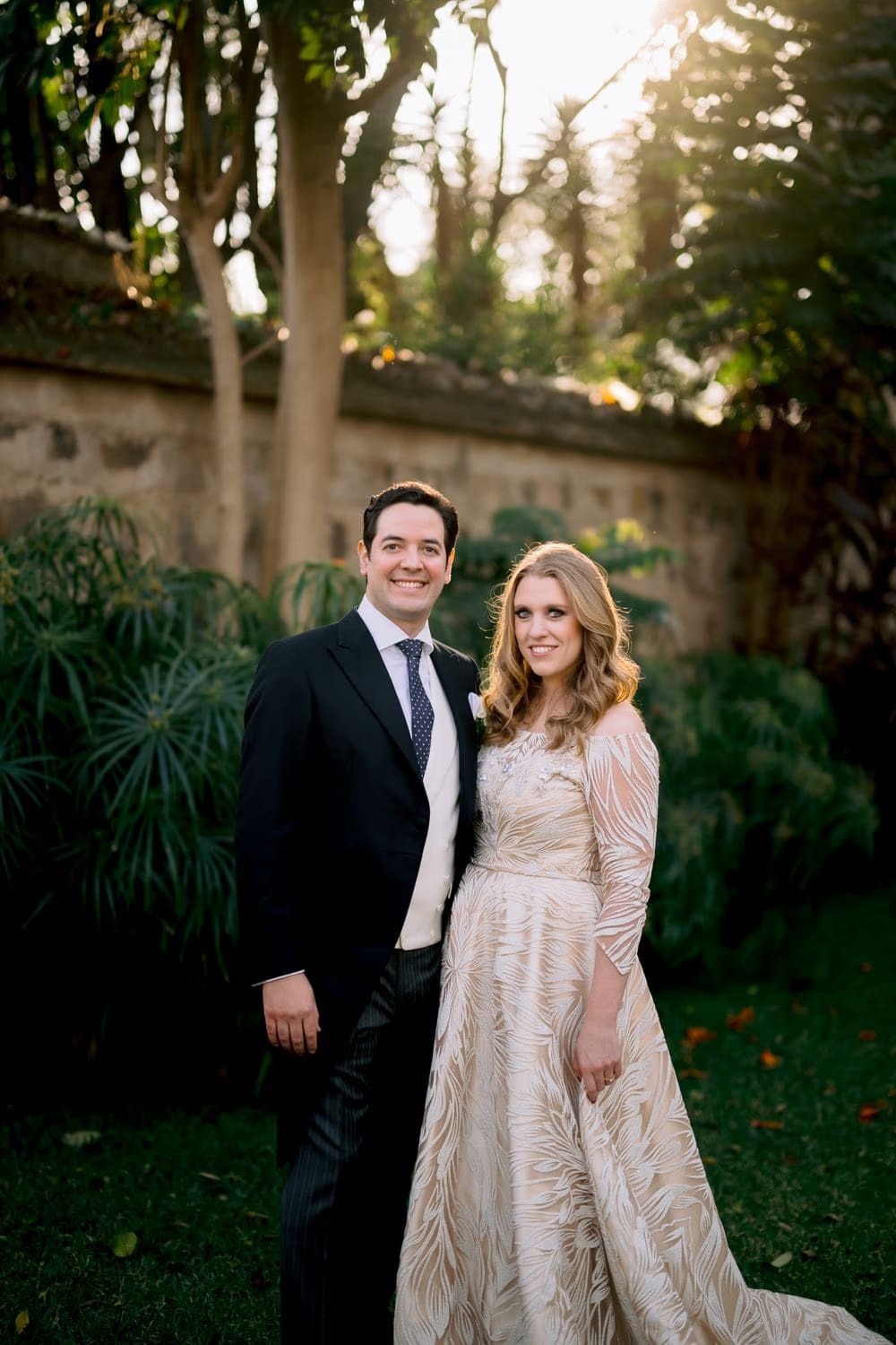 Wide couple portrait in a leafy walled garden at golden hour, with the bride's textured ivory gown catching the light — Antigua Guatemala wedding.