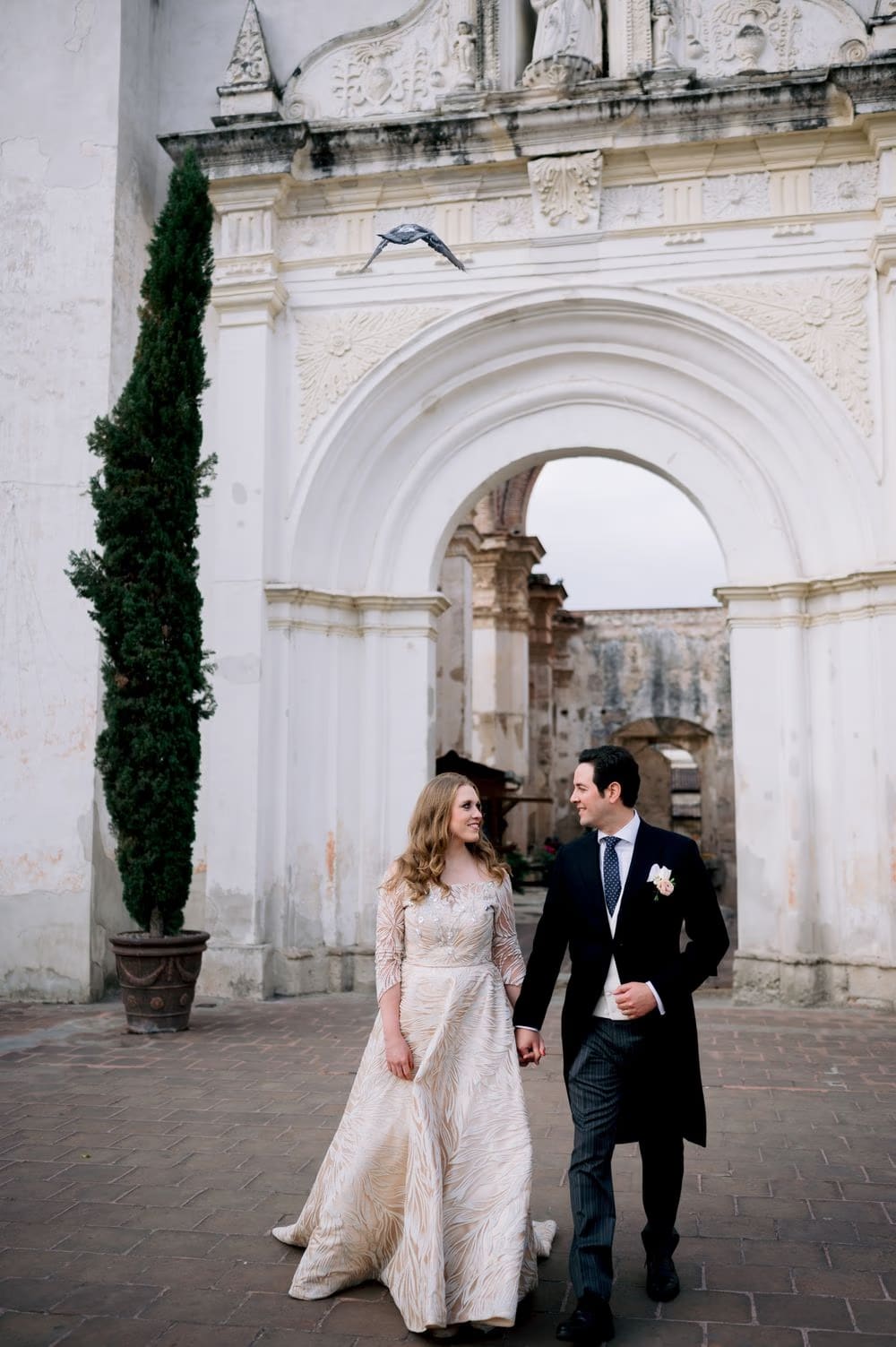Couple walk hand-in-hand under a tall white arch beside a slender cypress at a colonial ruins wedding in Antigua Guatemala.