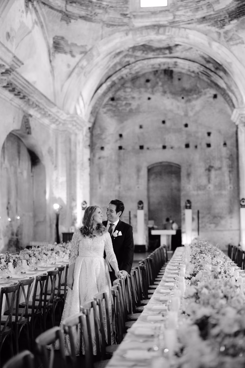 Black-and-white image of bride and groom embracing in the candle-lit nave of San José el Viejo Ruins, between long reception tables.