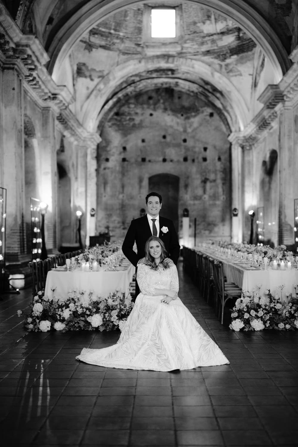 Bride seated in her ivory gown with the groom standing behind, framed by the vast vaulted nave of San José el Viejo Ruins — black-and-white editorial portrait.