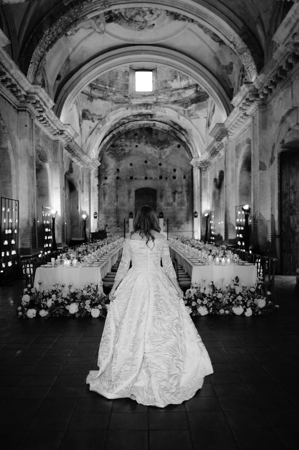 Bride seen from behind in her embroidered gown, gazing toward the candle-lit altar between two long banquet tables — intimate wedding in colonial ruins.