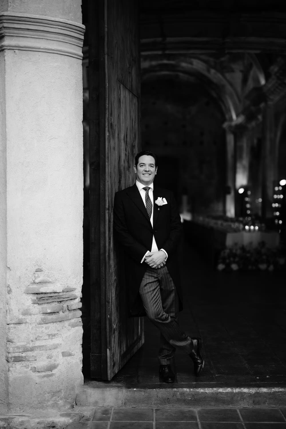Black-and-white groom portrait leaning against a column at the open doorway of the ruins — Antigua Guatemala wedding.