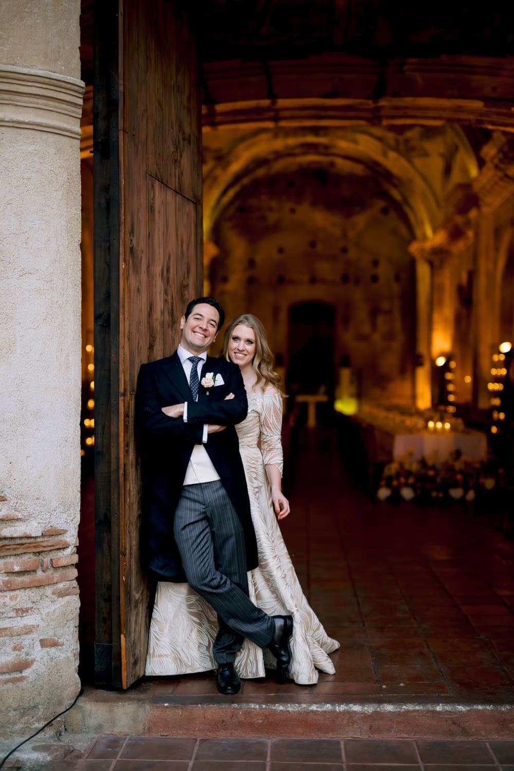 Bride and groom lean side-by-side against the giant wooden door of the ruins, candle-lit nave glowing behind them at their colonial Antigua wedding.