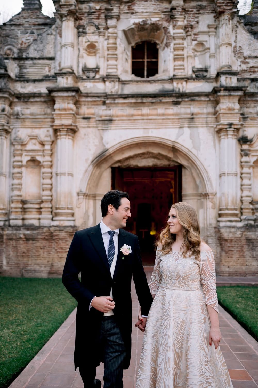 Couple walk hand-in-hand down the brick path leading away from the carved baroque entrance to the ruins in Antigua Guatemala.