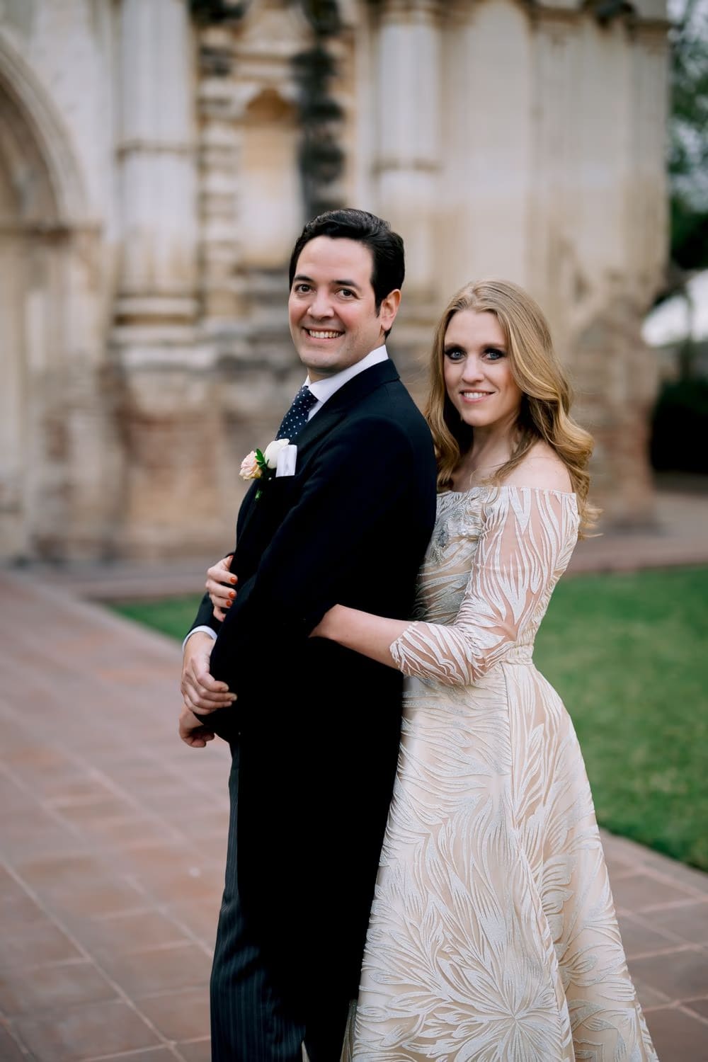 Bride wraps her arms around the groom from behind, both smiling at the camera in front of the weathered colonial facade — colonial Antigua wedding.