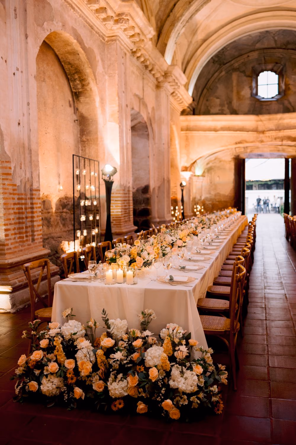 Long reception table with cascading peach and white florals by Cynthia Carrillo, taper candles glowing against the brick arches of San José el Viejo Ruins.