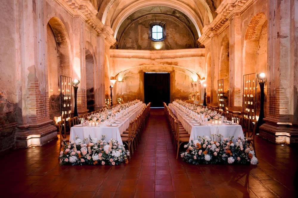 Symmetrical view down the central aisle between two banquet tables framed by floral bases and votive candles — intimate wedding reception in the ruins.