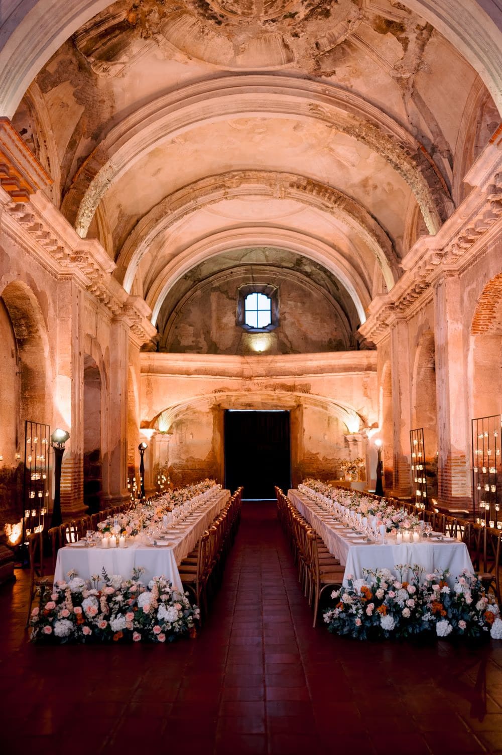 Wide vertical view of the empty nave of San José el Viejo Ruins set up for the reception, with two long tables flanked by floral installations and candle towers.