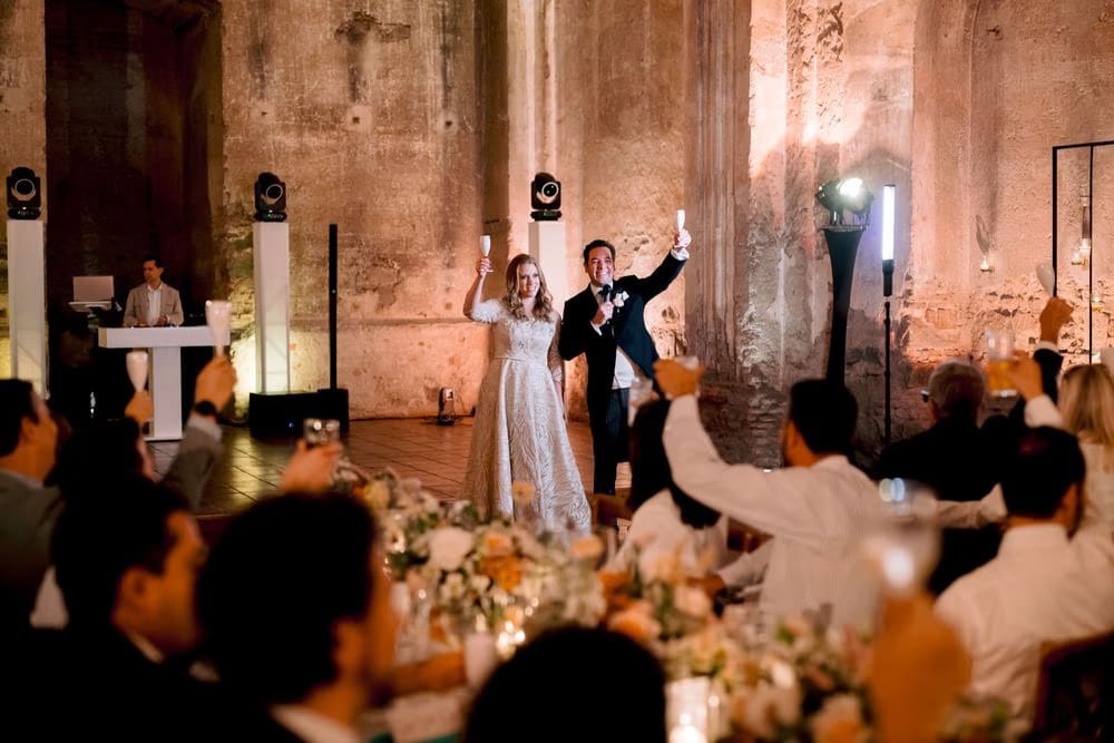 Bride and groom raise their champagne flutes for a toast as guests cheer at the candle-lit reception inside the ruins.