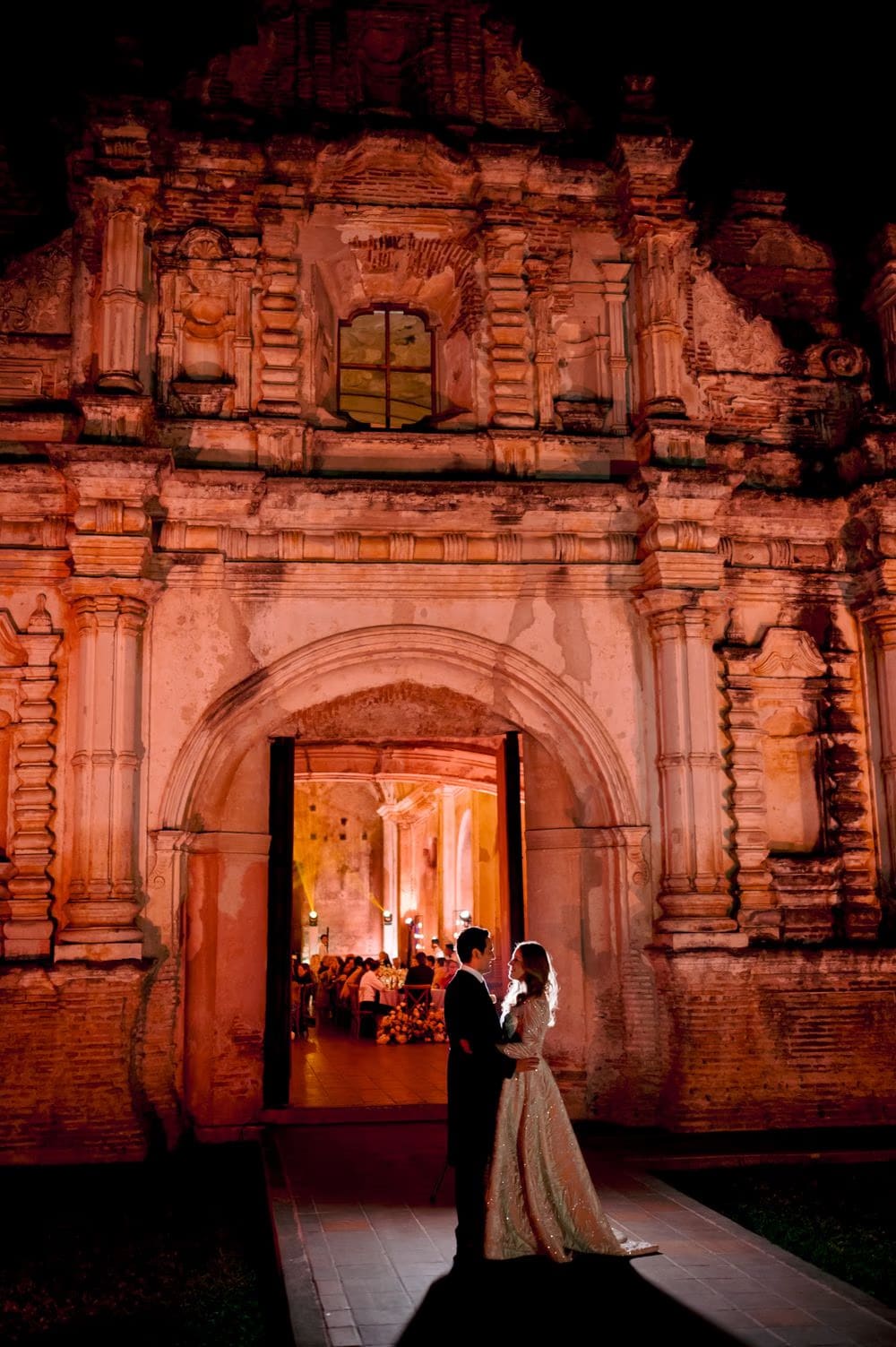 Ana and Jose embrace under the warm-lit baroque facade of the ruins at night, the glowing reception visible through the open archway.