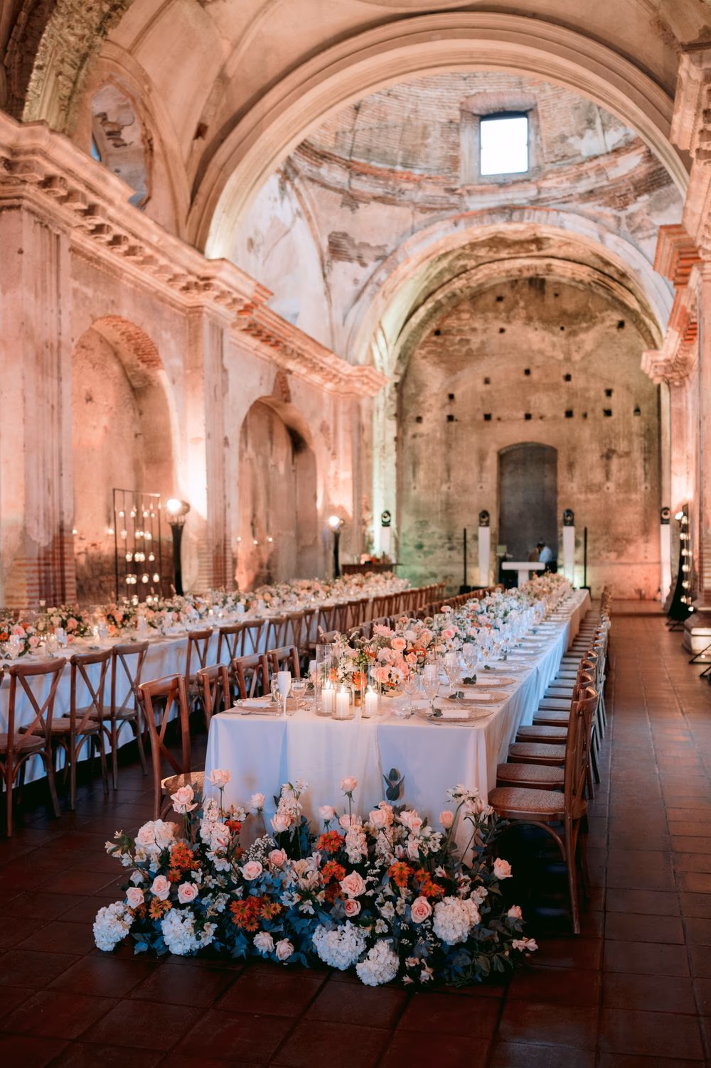 Long candle-lit reception table runs through the vaulted nave of San José el Viejo Ruins with a lush floral installation in the foreground — Ana and Jose wedding in Antigua Guatemala.
