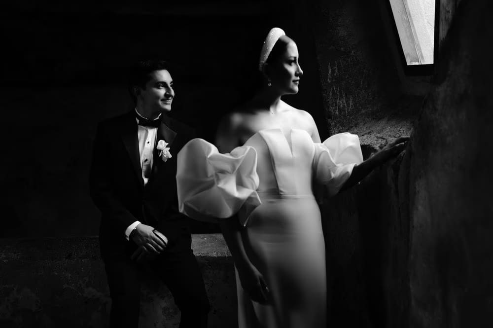 Groom seated beside bride leaning at a stone window inside Capuchinas Ruins — moody black-and-white editorial portrait.