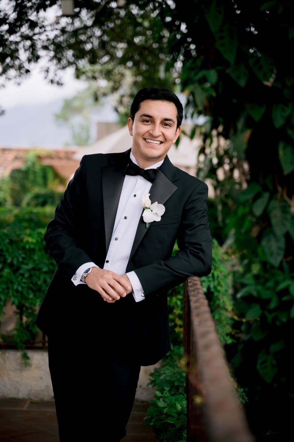 Groom Josue in a black tuxedo with white orchid boutonniere leans against a railing during getting-ready in Antigua Guatemala.