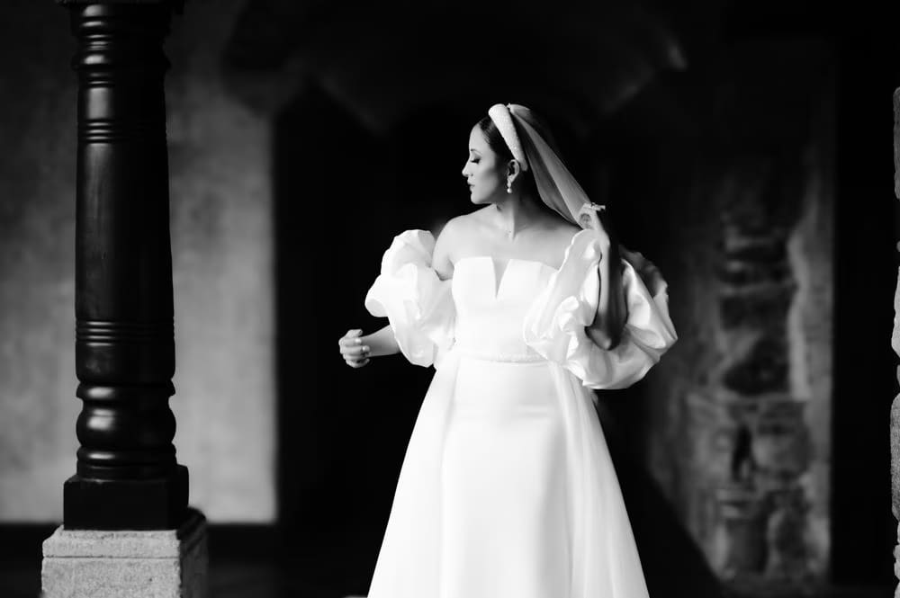 Bride in puff-sleeve gown and veil stands among black stone columns in a corridor of the convent ruins — editorial black-and-white.