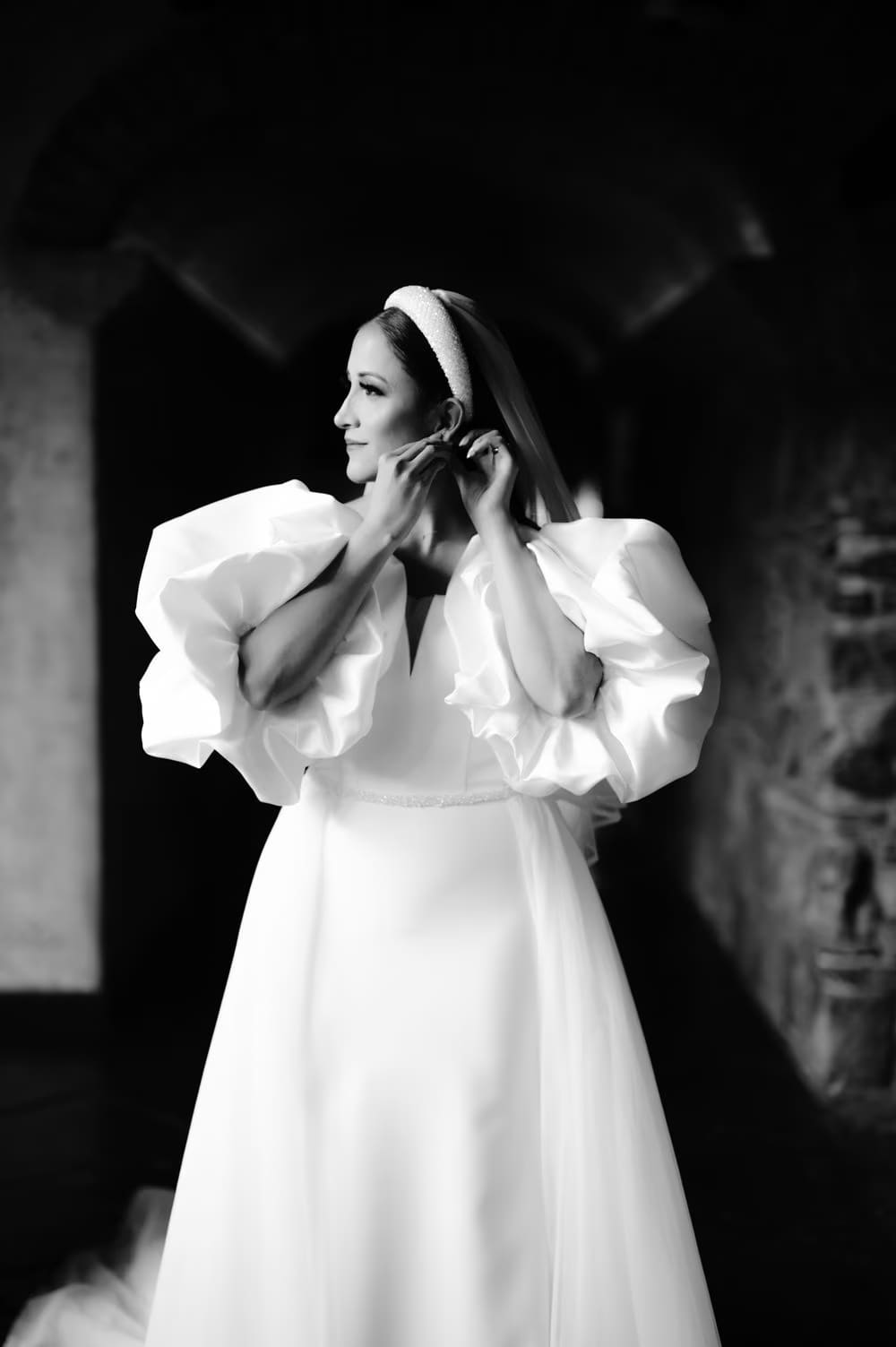 Bride adjusts her earring in a dim stone passage — candid getting-ready moment captured in black-and-white.