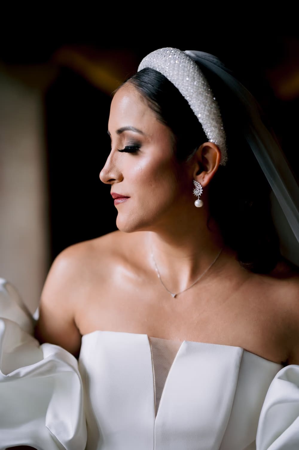 Close bridal portrait — soft side light, pearl drop earring and crystal headband against a dark backdrop.