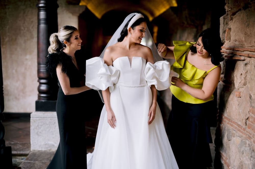 Mother and friend help the bride into her puff-sleeve wedding gown in a colonial corridor of Antigua Guatemala.