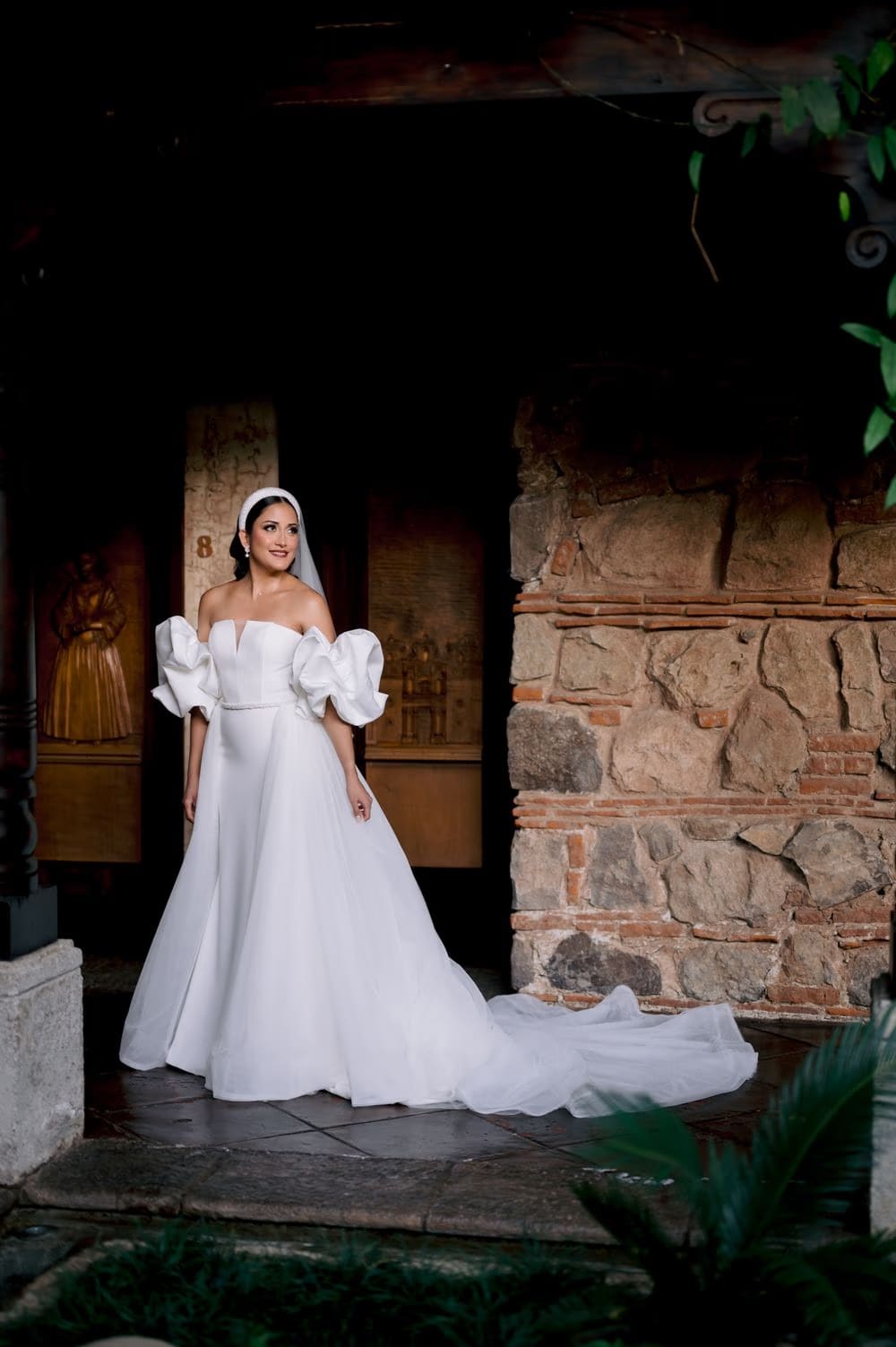 Full-length bridal portrait — train spread on the tile floor framed by a stone arch and brick wall.