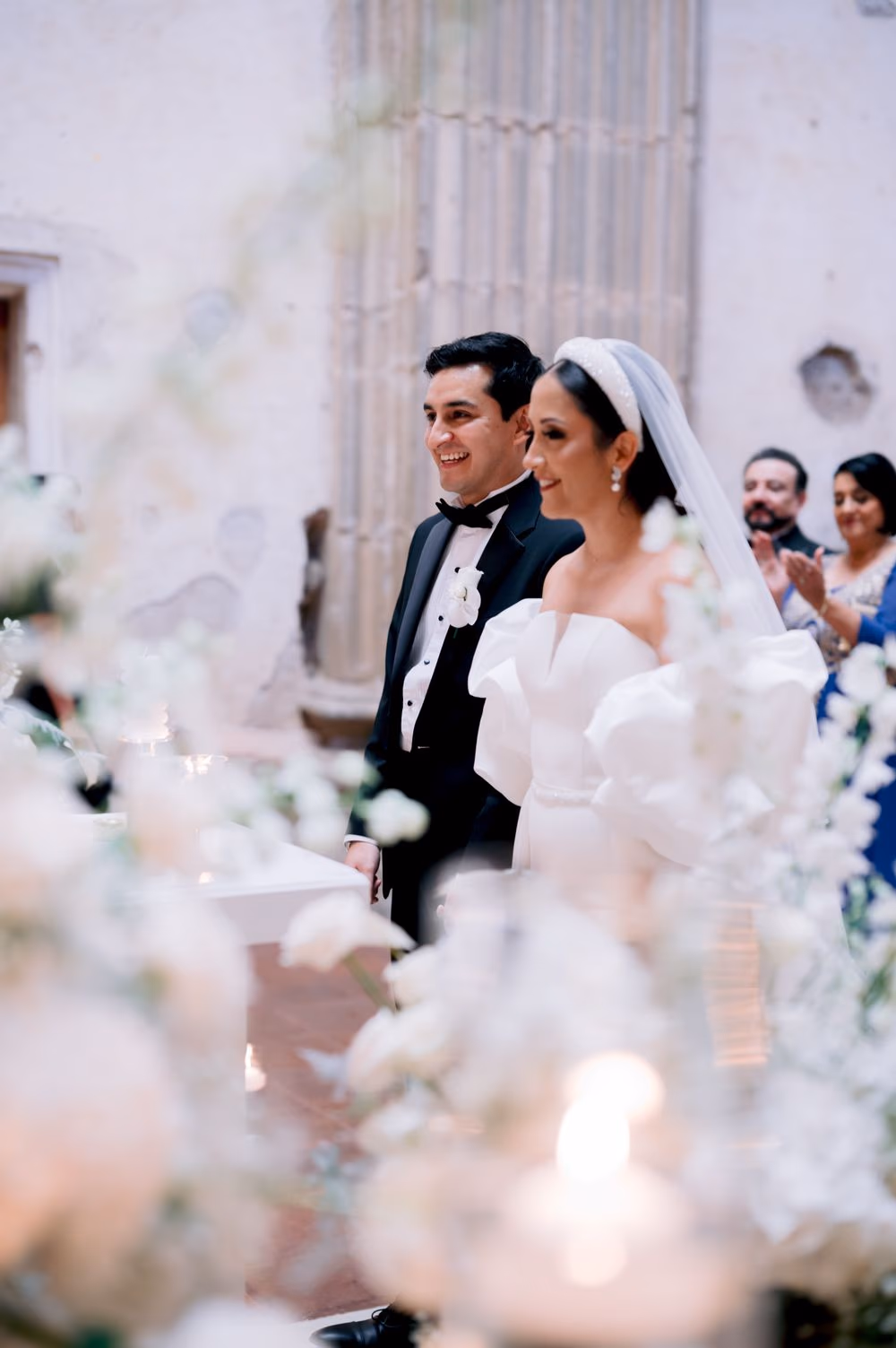 Bride and groom share a beaming smile at the ceremony framed by white florals at Capuchinas Ruins, Antigua Guatemala.