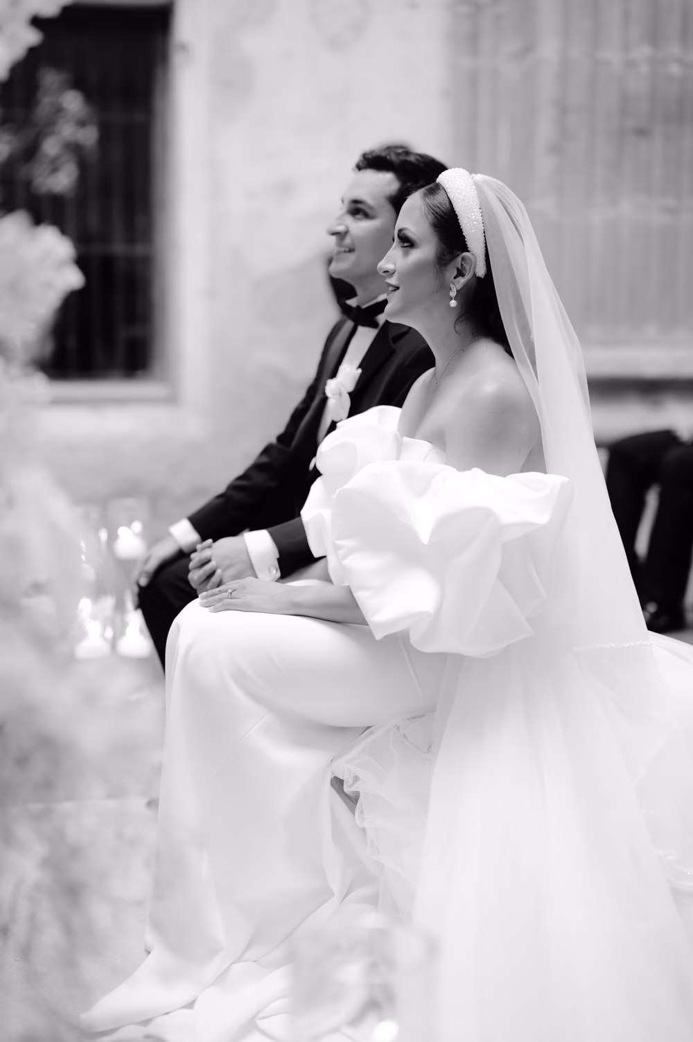Bride and groom seated side by side during the religious ceremony, tied with the lazo cord — black-and-white.