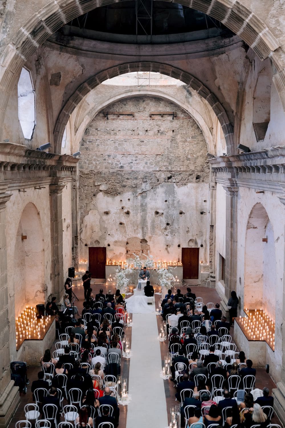 Wide elevated view of the wedding ceremony inside the soaring stone arches of Capuchinas Ruins, candle-lit aisle and seated guests.
