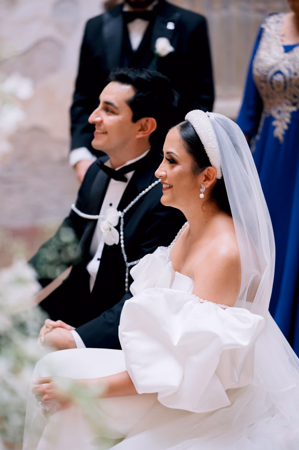 Bride and groom sit together during the ceremony with parents standing behind — black-and-tie intimate detail.