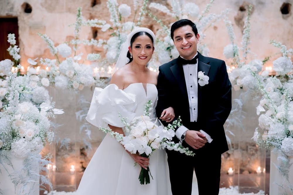Bride and groom pose smiling at the white floral altar framed by tapered candles.