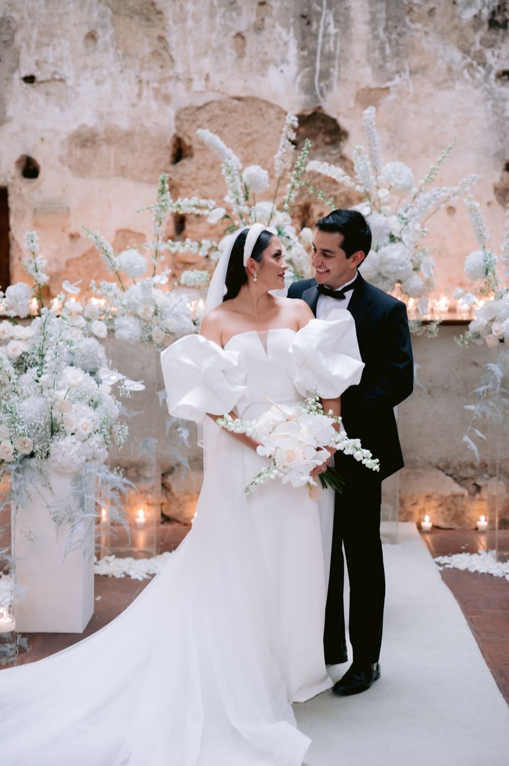 Bride and groom share a look beside an extravagant white floral arch inside Capuchinas Ruins — editorial wedding portrait.