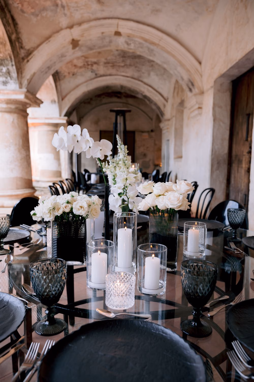 Wide reception table under vaulted arches set with white orchids, snapdragons and pillar candles — decor by Decor Guatemala.