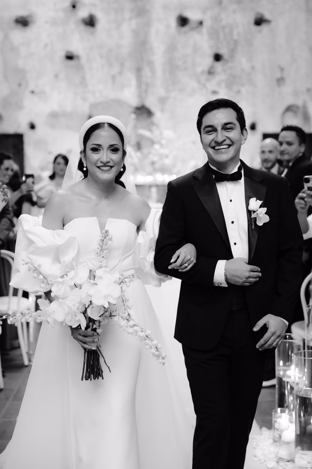 Bride and groom beam as they walk back down the aisle after the ceremony, surrounded by applauding guests — black-and-white.
