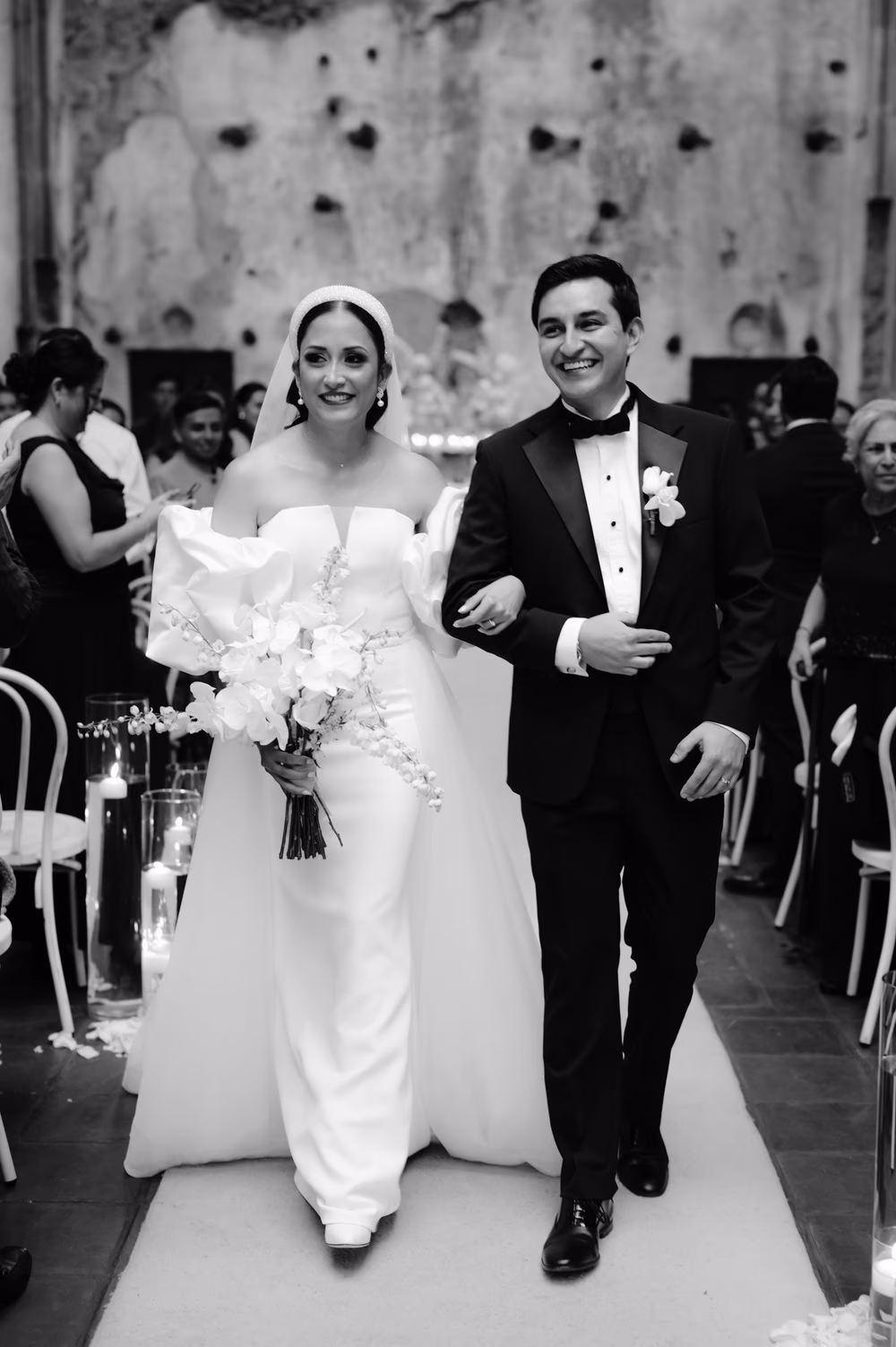 Newlyweds walk the aisle arm in arm carrying a cascade orchid bouquet — black-and-white editorial frame.
