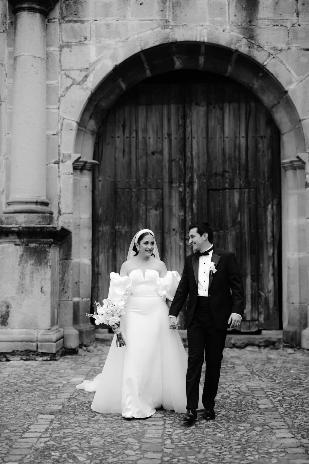 Bride and groom walk hand in hand on cobblestones in front of the convent's wooden colonial doors — destination wedding portrait.