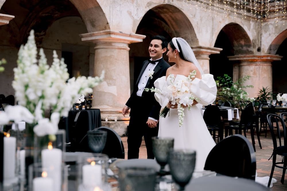 Newlyweds laugh as they enter the cloister reception lit with string lights and lined with white floral towers.
