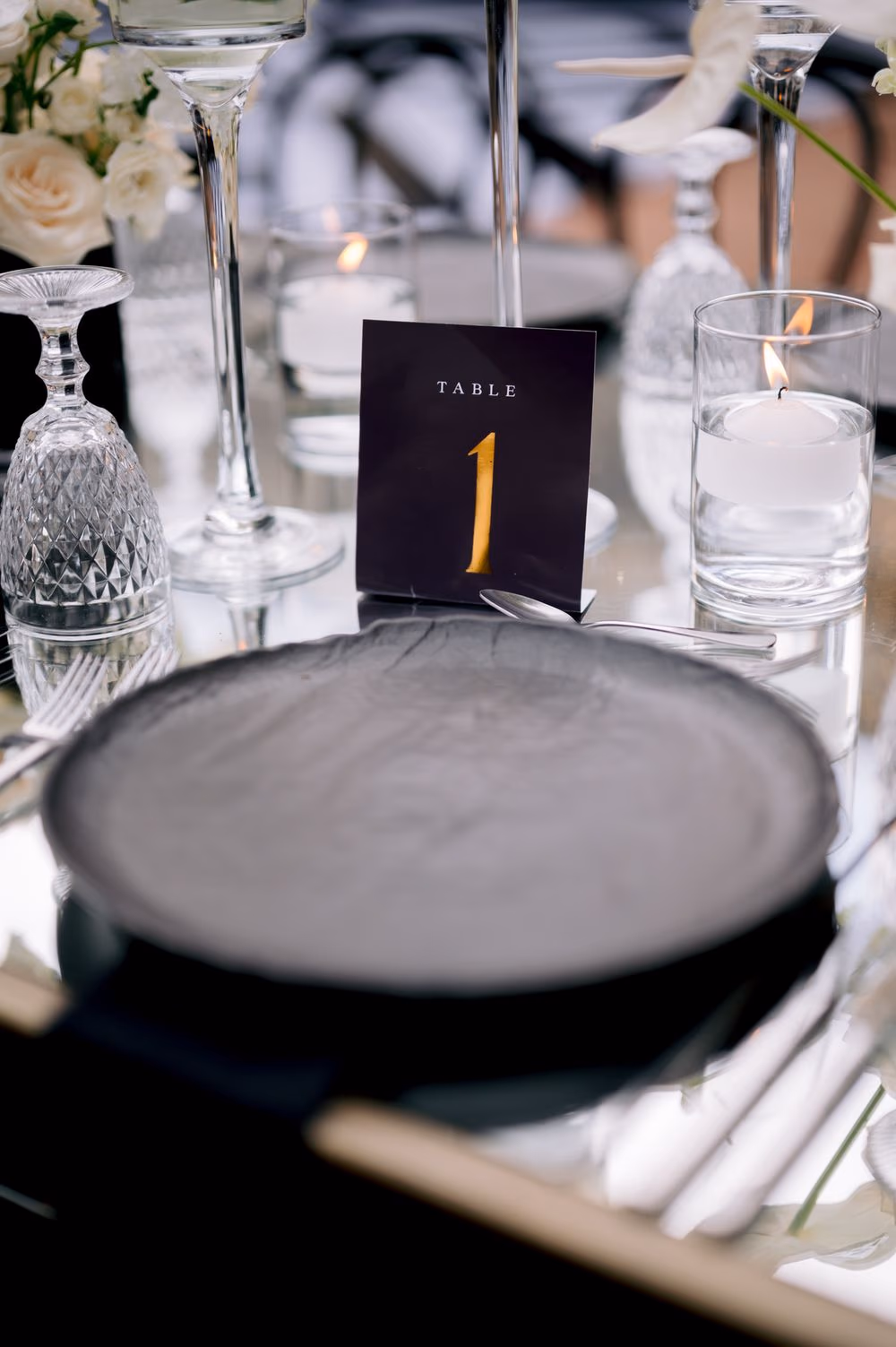 Table number card in deep navy with a gilded numeral one, surrounded by stemware and floating candles.